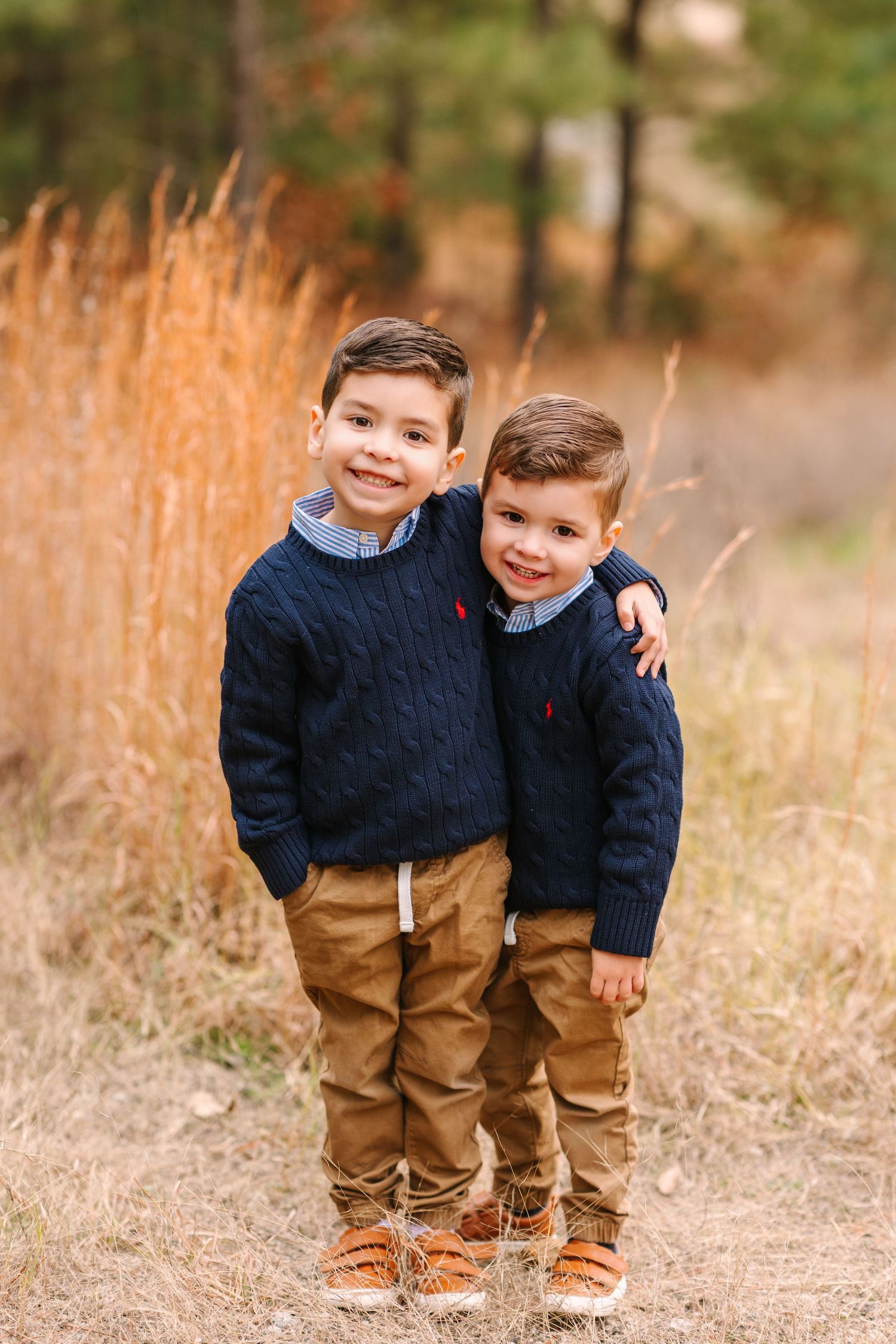 Two young children in matching navy sweaters and khaki pants stand together in a field of tall grass at sunset.