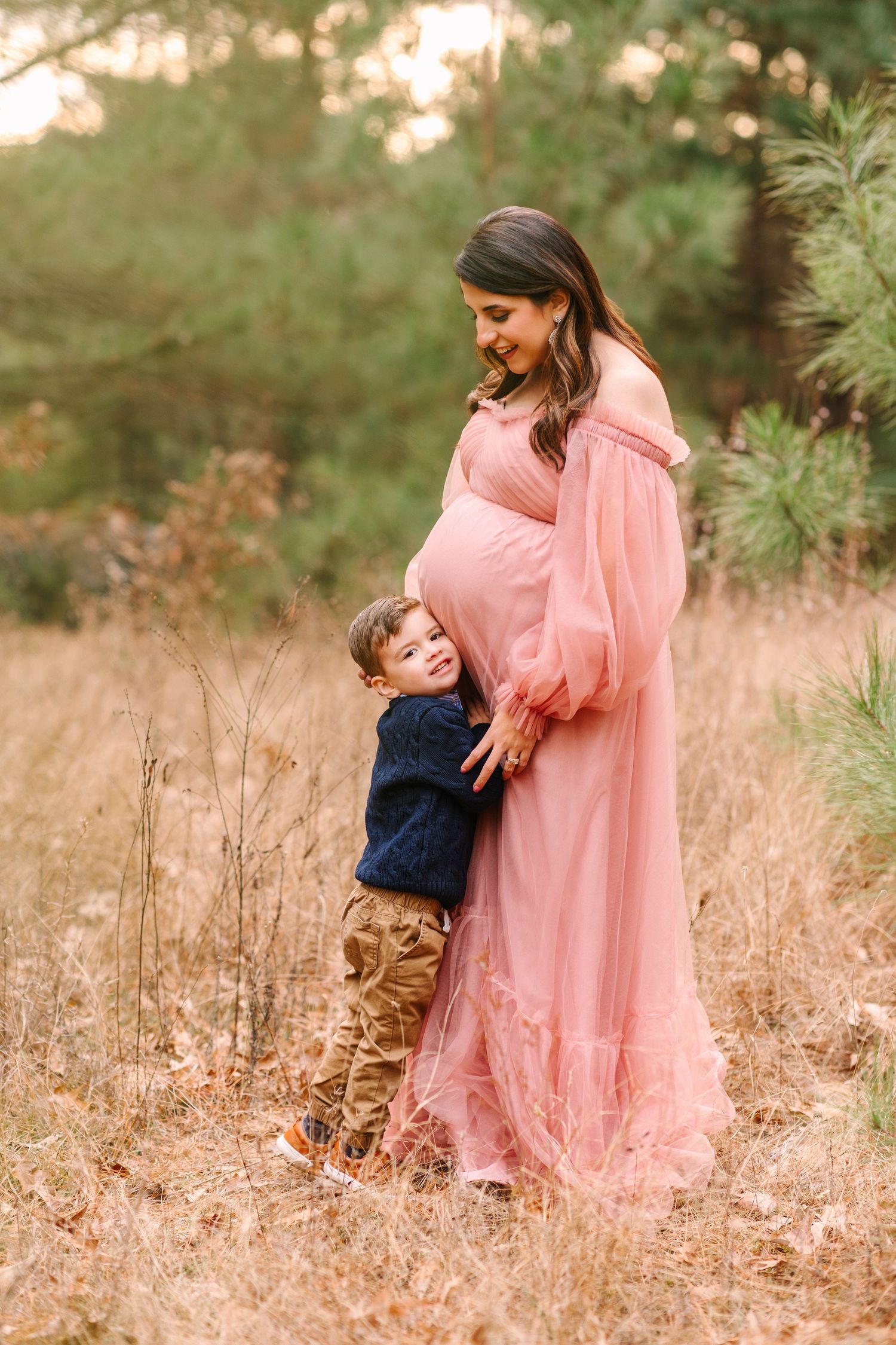 A pregnant woman in a flowing pink dress shares a tender moment with a young child in a rustic outdoor setting.
