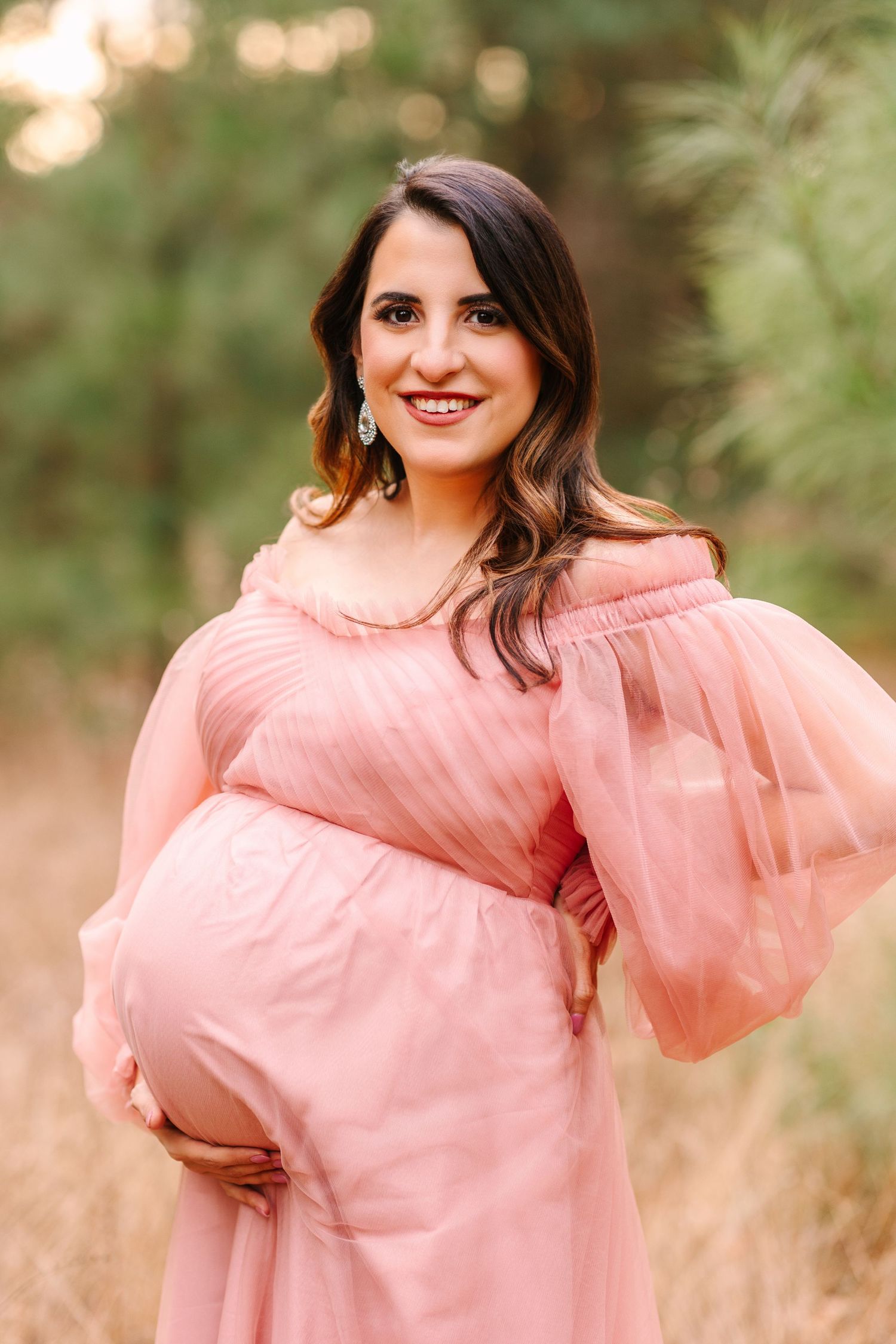 A pregnant woman wearing a pink off-shoulder dress poses outdoors during golden hour.