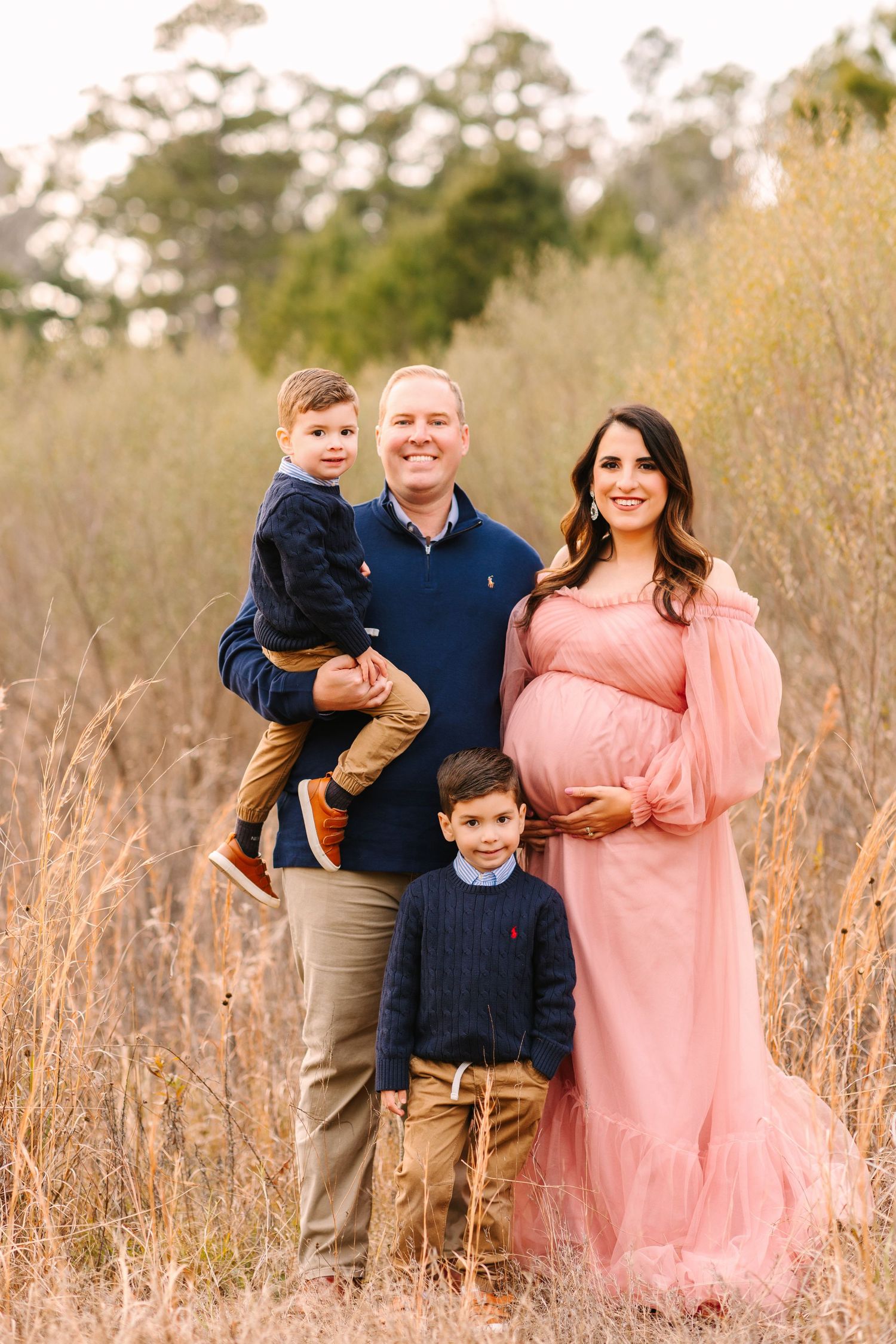 Family of four poses together in a field with dried grass, mom wearing a pink maternity dress.