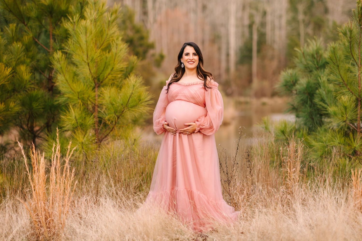 A woman in a flowing pink dress poses elegantly in a natural outdoor setting with pine trees in the background.
