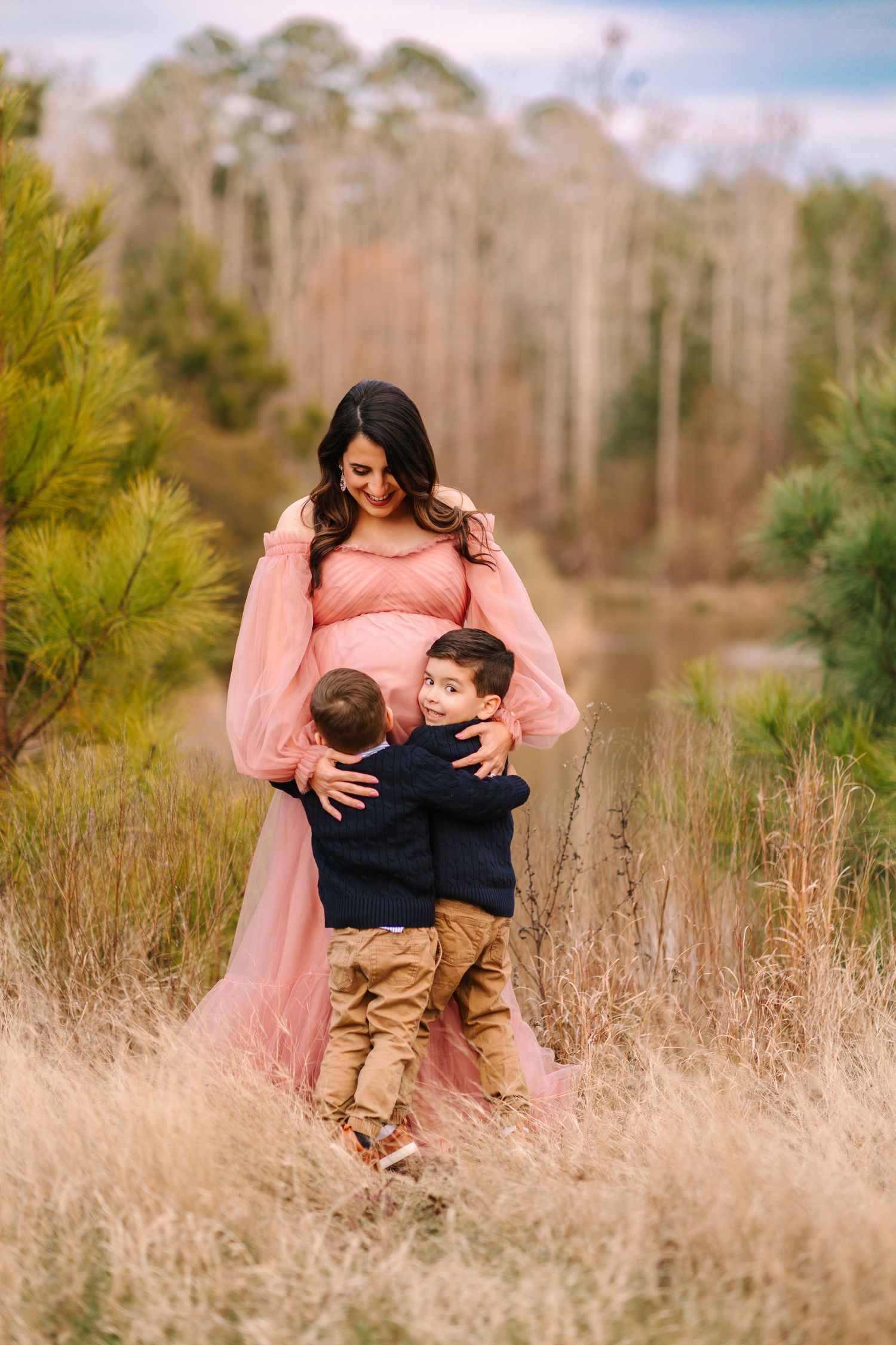 Woman in pink dress hugs two children while standing in field of tall grass.