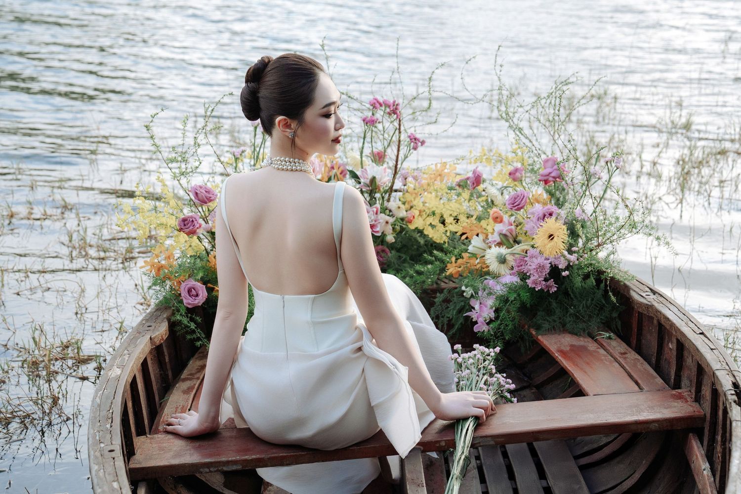Graceful bride in backless white dress sitting in a wooden boat surrounded by colorful flowers.