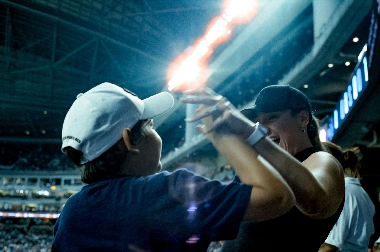 Baseball fans raise their arms in unison while watching a night game at a crowded stadium.