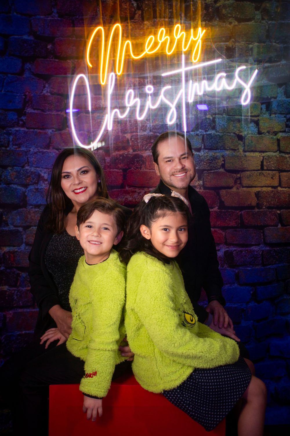 Children in neon yellow sweaters pose against a brick wall with illuminated neon Christmas signs.