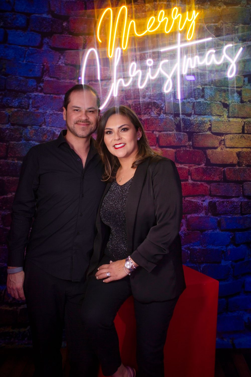 A couple poses together in front of a brick wall illuminated by colorful neon signs in what appears to be a nightclub setting.