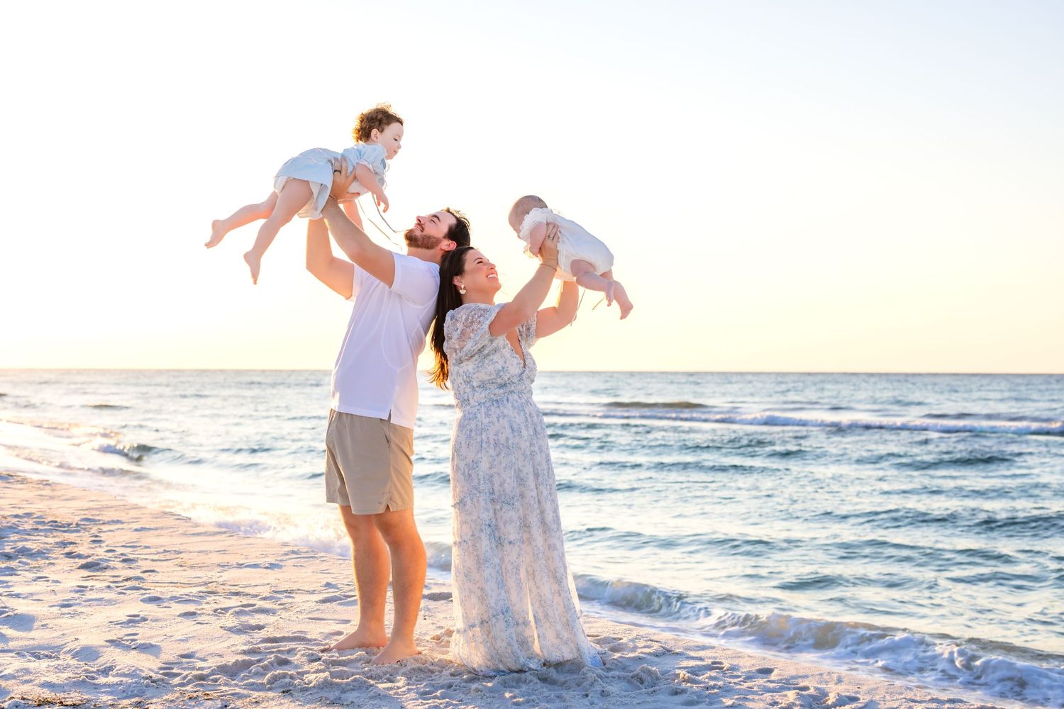 Family enjoying playful moments together on a sunny beach at sunset with ocean waves in the background.