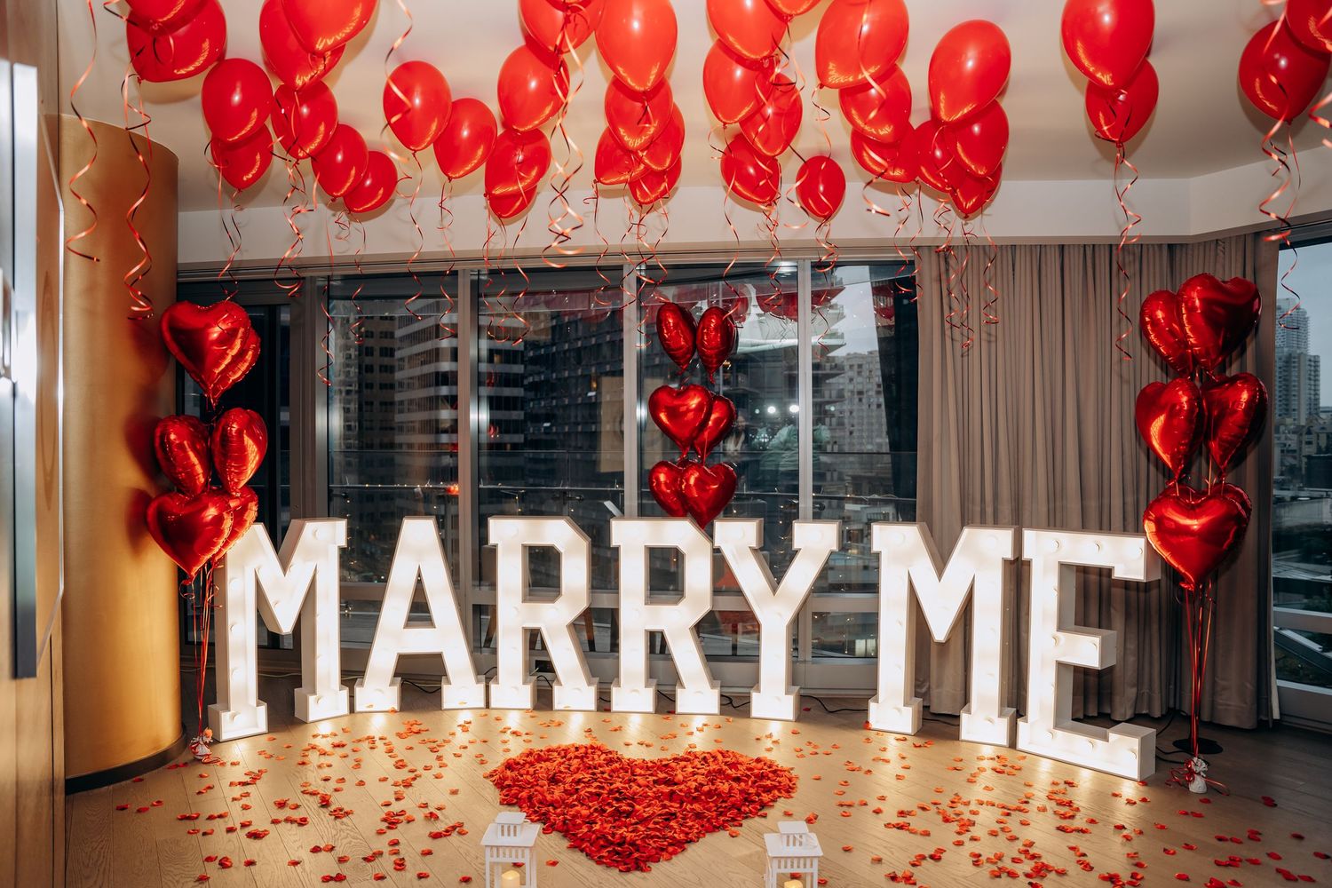Red heart balloons and rose petals decorate a room with 'MARRY ME' text display for a proposal.