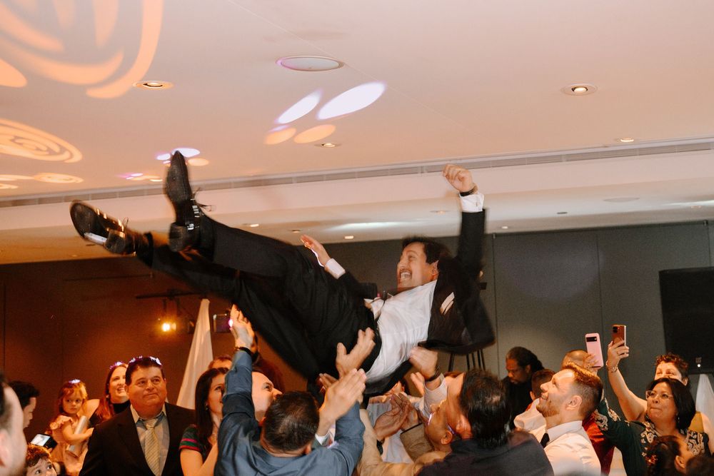 Groom being lifted by crowd during lively wedding reception celebration indoors.