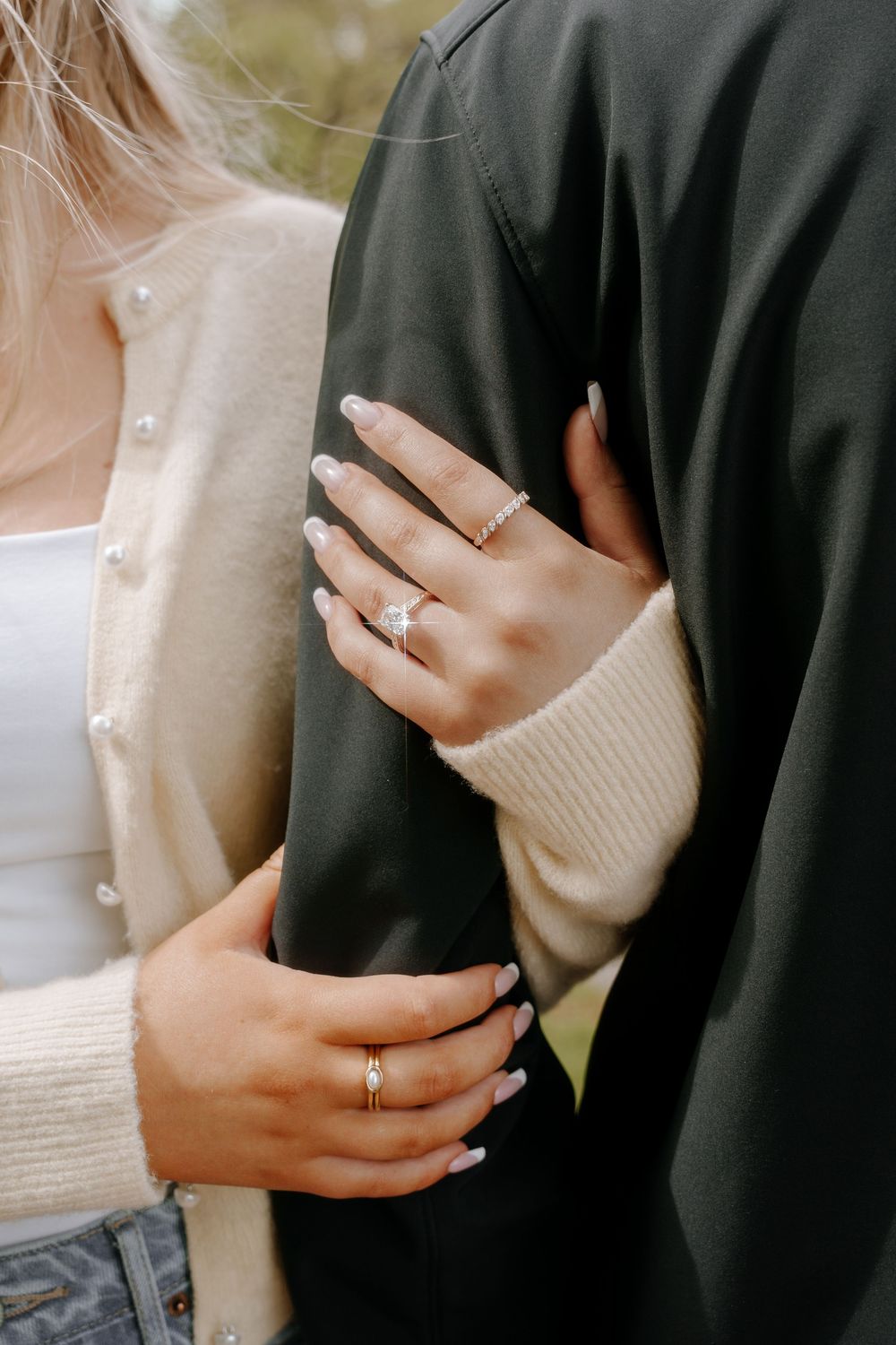Close-up of a woman's hand with an engagement ring and gold band, embracing a man in a dark jacket.