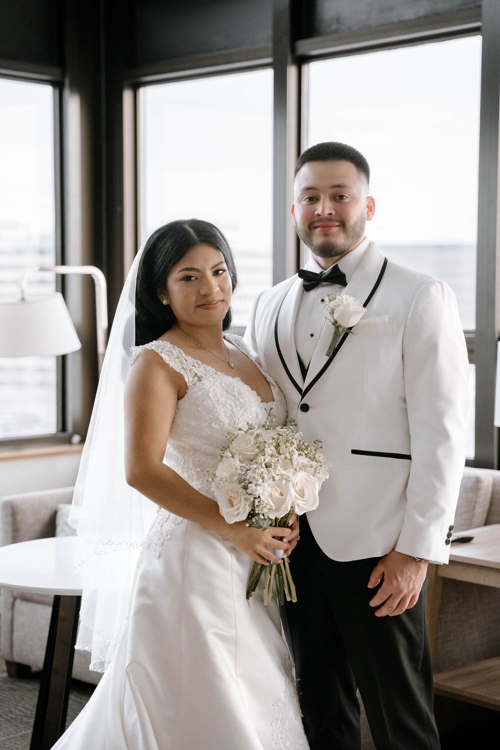 Bride in lace gown holding white bouquet stands with groom in white tuxedo jacket indoors.