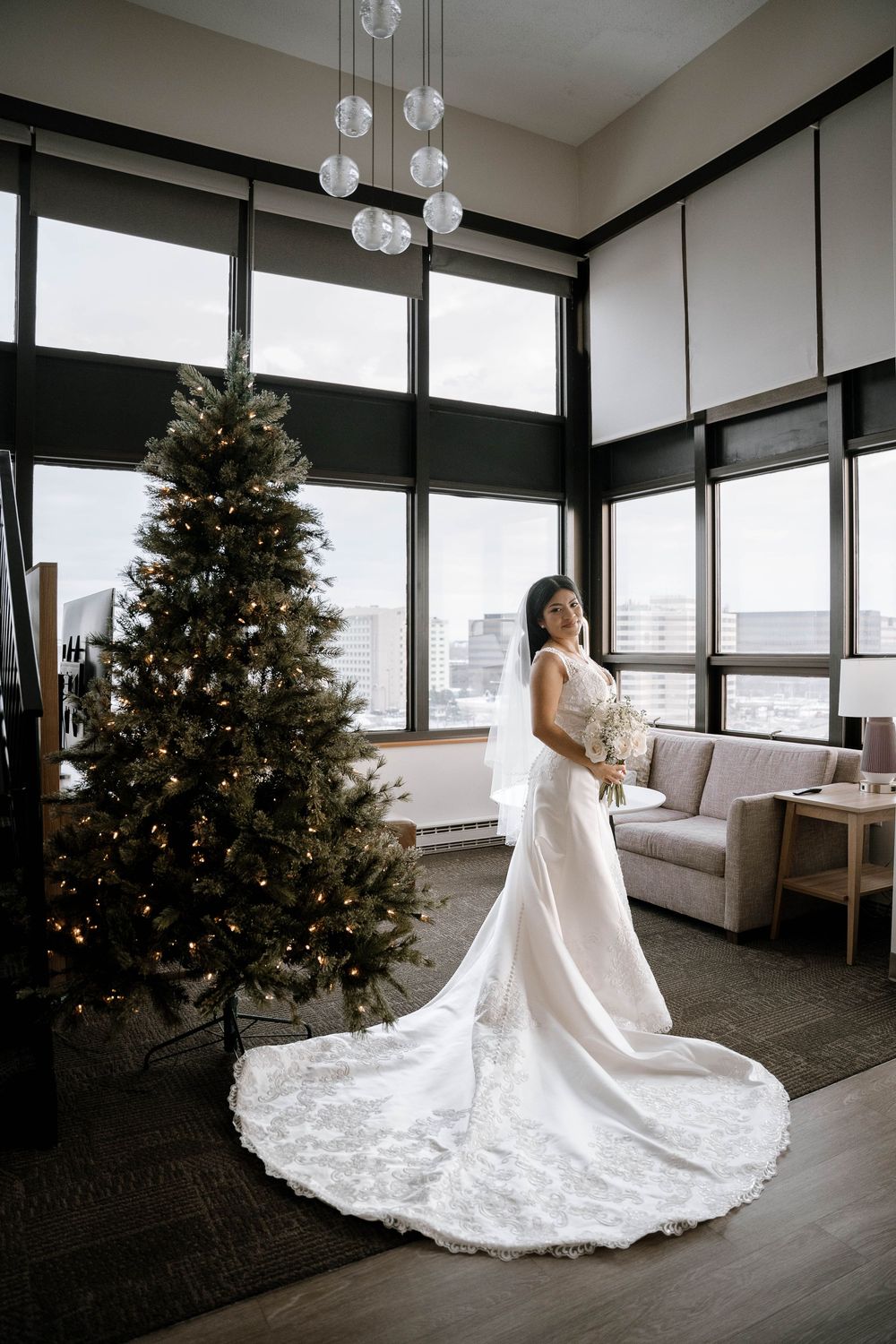 Bride in elegant white gown with lace train stands beside decorated Christmas tree in modern hotel suite.