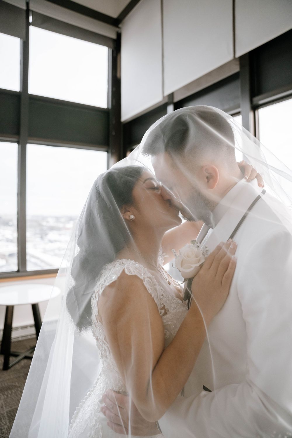 Bride and groom sharing a romantic kiss under a white veil near large windows in an elegant indoor wedding setting.
