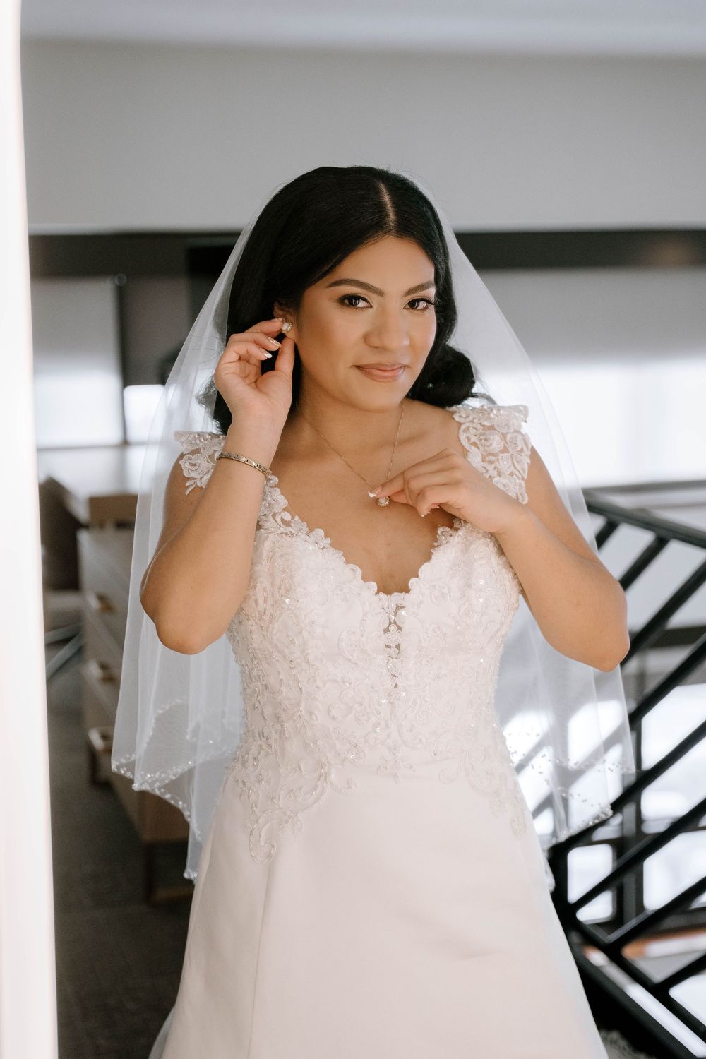 Bride in white lace wedding gown and veil adjusting earring, smiling elegantly indoors.
