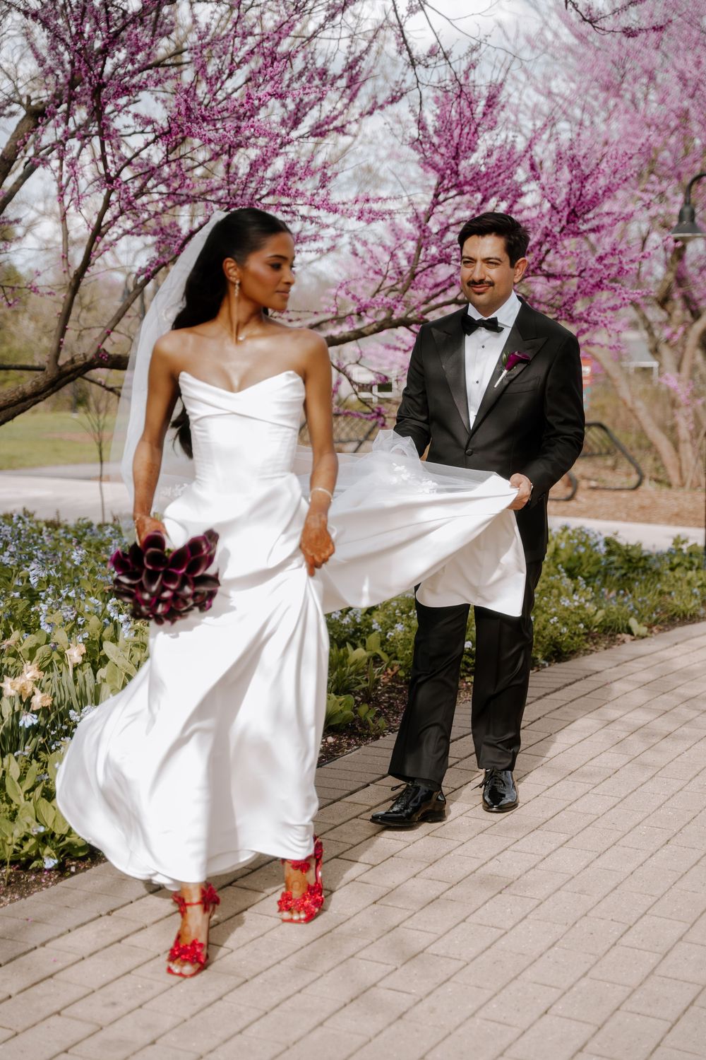 Bride in white strapless gown with red heels walks ahead of groom in tuxedo near blooming pink trees.