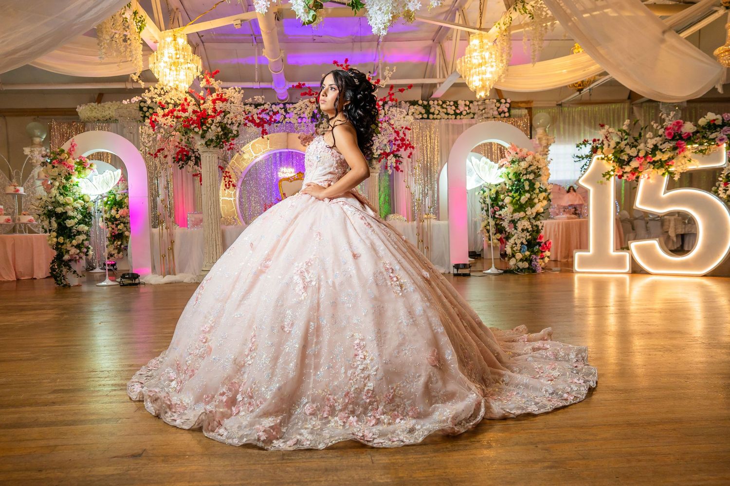 A grand ballroom decorated for a quinceañera celebration with pink lighting and floral arrangements.