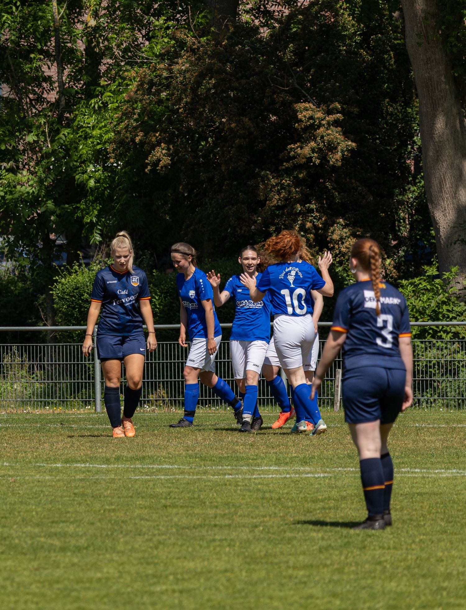 Women's soccer match in progress with players in blue and navy uniforms competing on a sunny grass field.