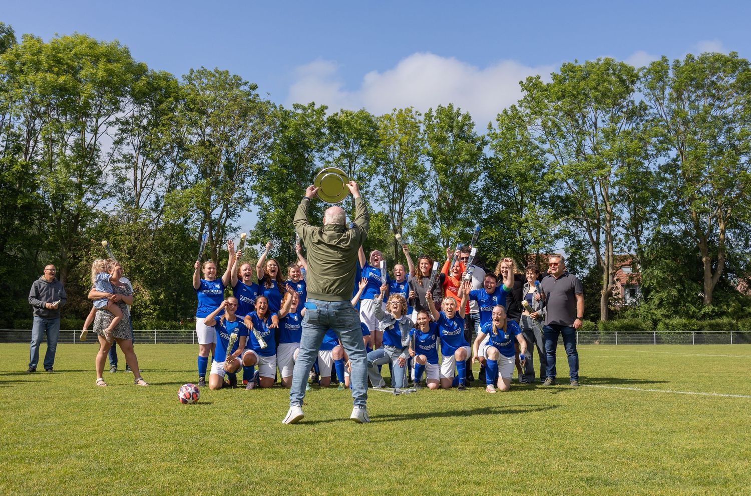 A group of sports team players gathered on a sunny grassy field with trees in the background.