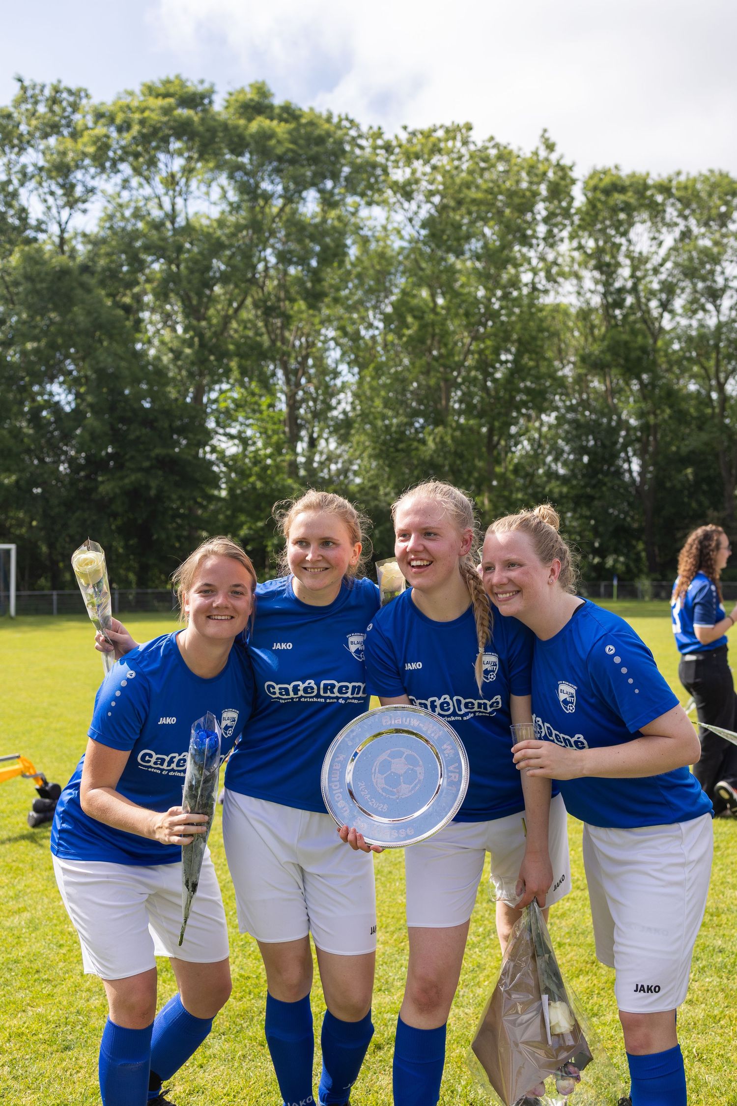 Athletes in blue and white uniforms posing together with trophies on a grass field with trees behind them.