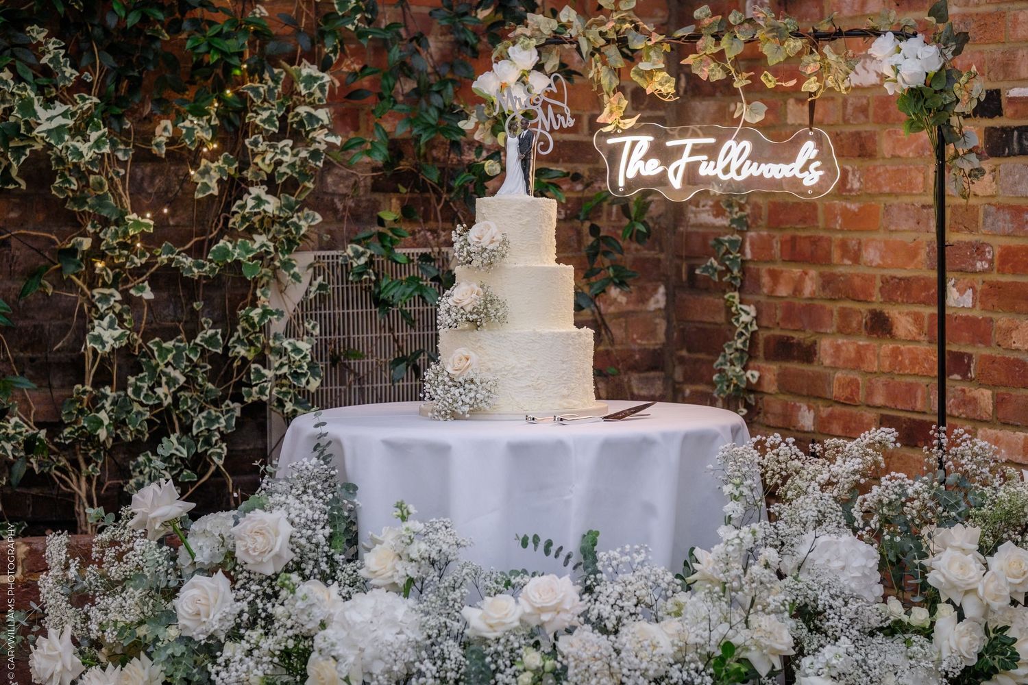White wedding cake decorated with delicate baby's breath flowers sits elegantly against a rustic brick wall backdrop.