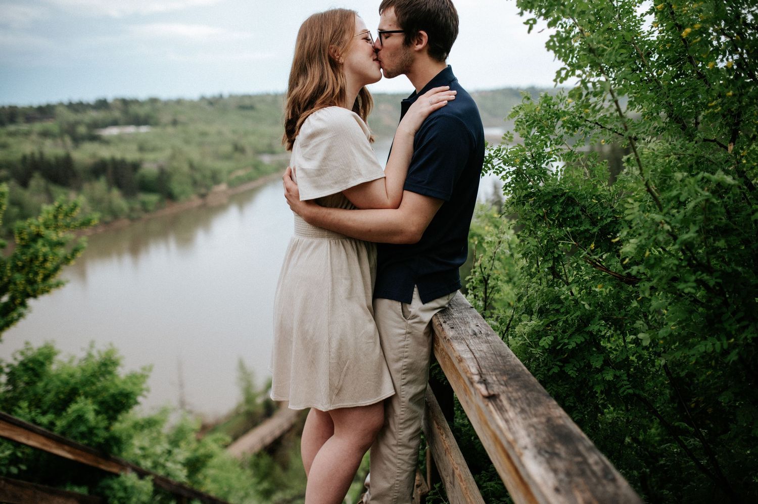 A couple embraces lovingly while standing on a path overlooking a scenic river bend in nature.
