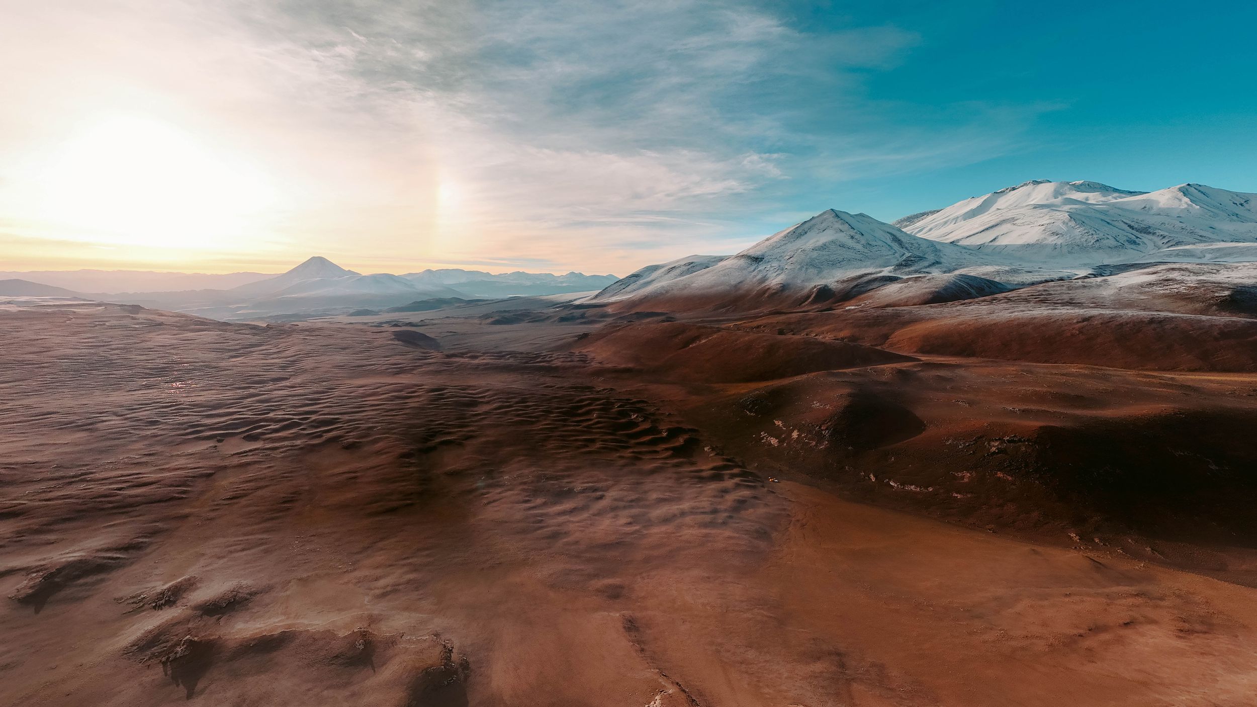 A stunning panoramic landscape of snow-capped mountains and desert terrain under a bright blue sky during sunset.