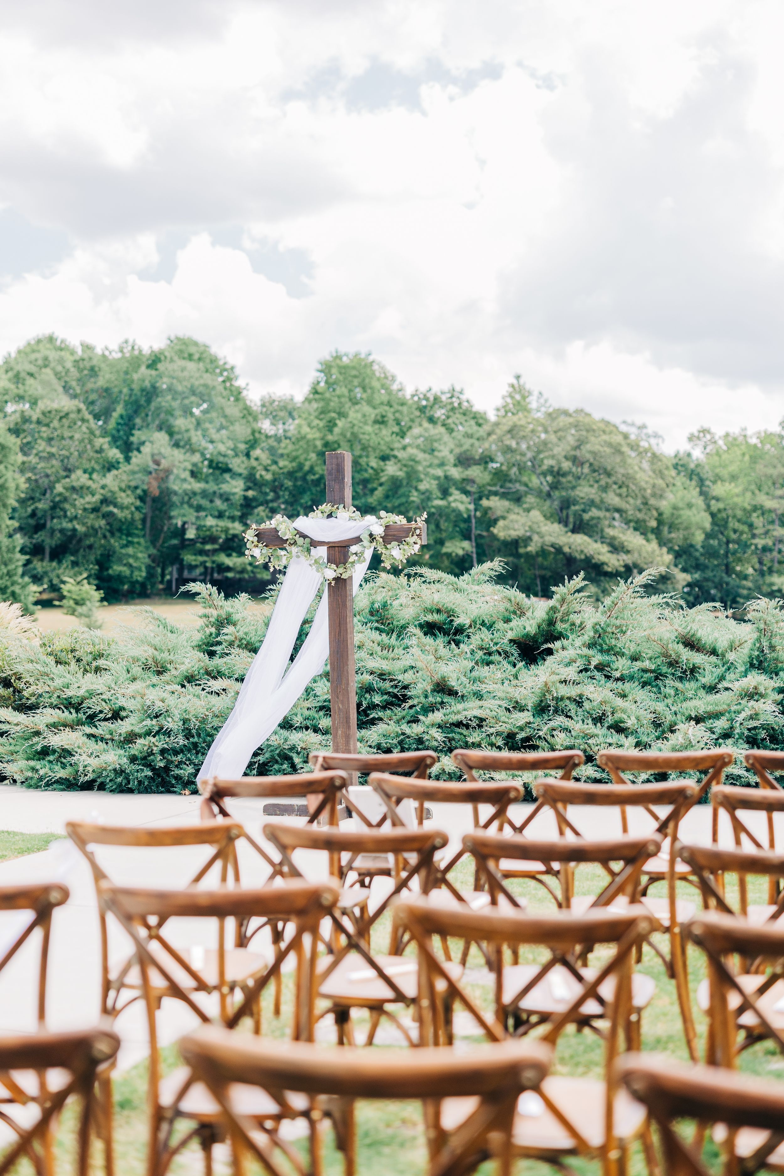 Rustic wooden cross-back chairs arranged outdoors for a garden ceremony with white fabric draping and greenery in background at Barn South