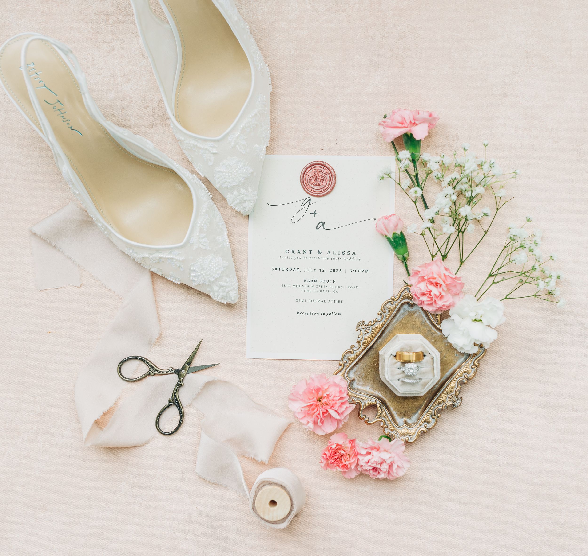 White wedding shoes, invitations, floral carnations, and vintage spools arranged in a soft, romantic flatlay composition.