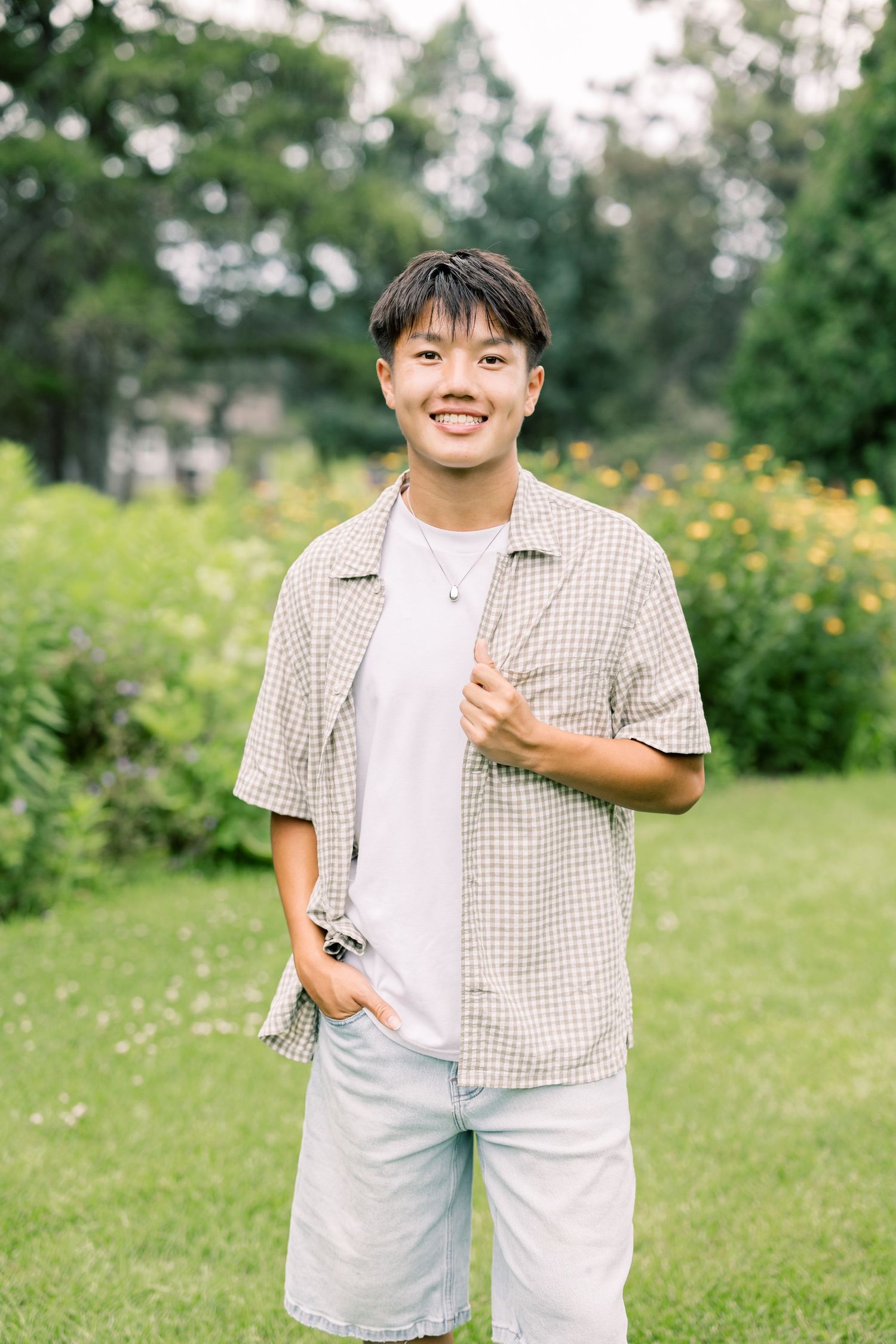 Person in a beige checkered shirt and white t-shirt stands outdoors in a sunny garden with yellow flowers.