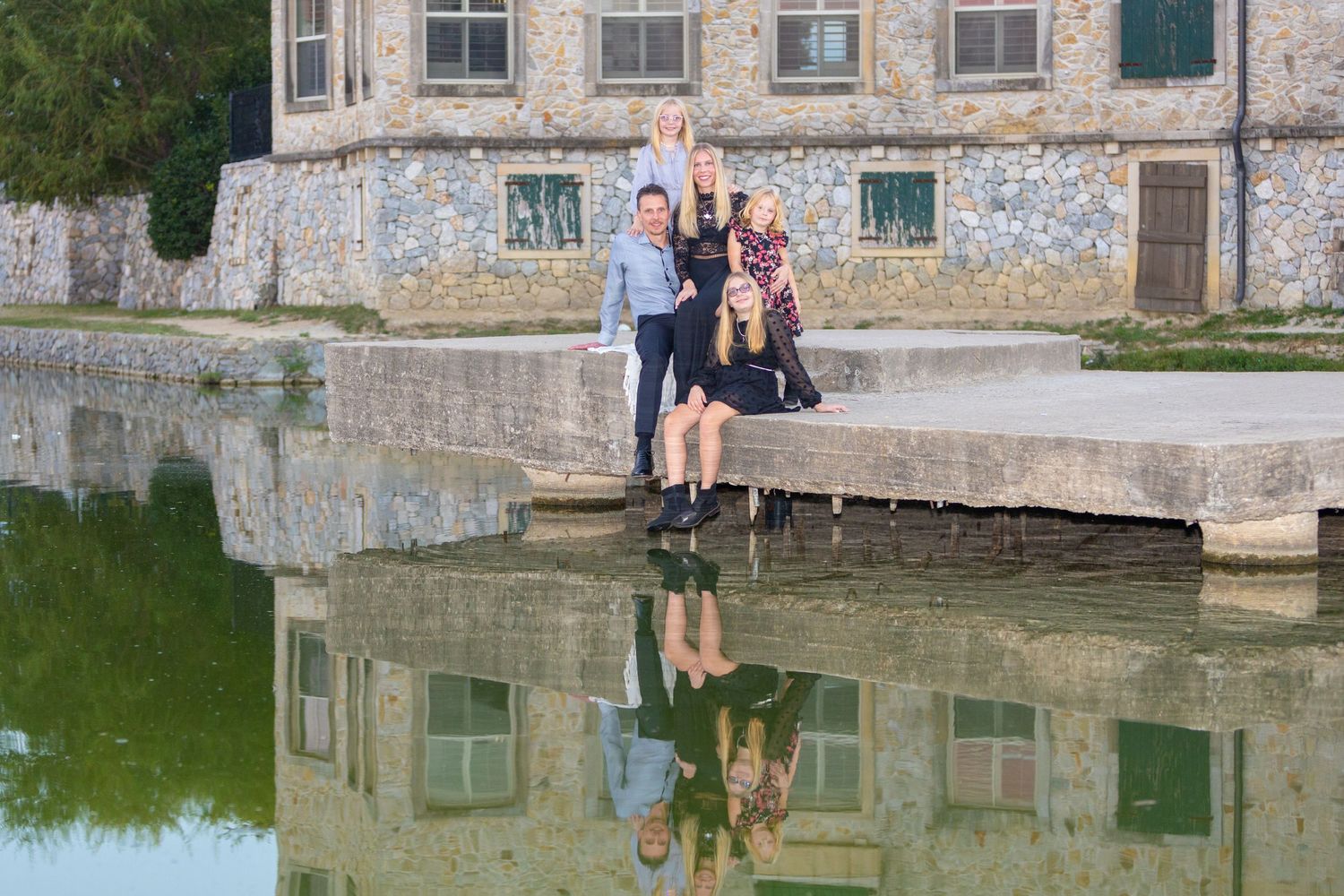 A series of stone walls and buildings reflected in calm water with people relaxing along the waterfront edge.
