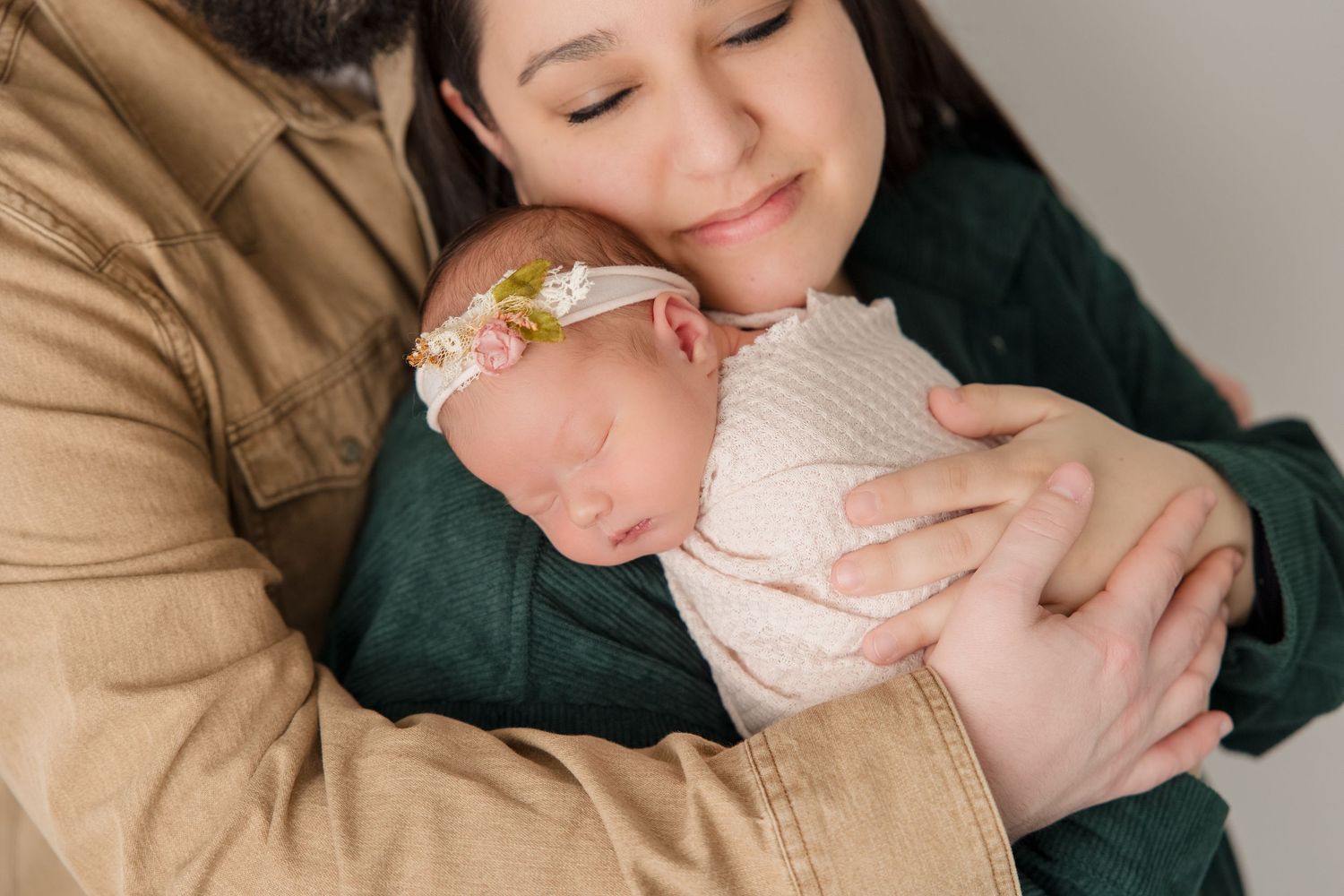 Tender parental embrace with sleeping newborn swaddled in soft white fabric against neutral backdrop.