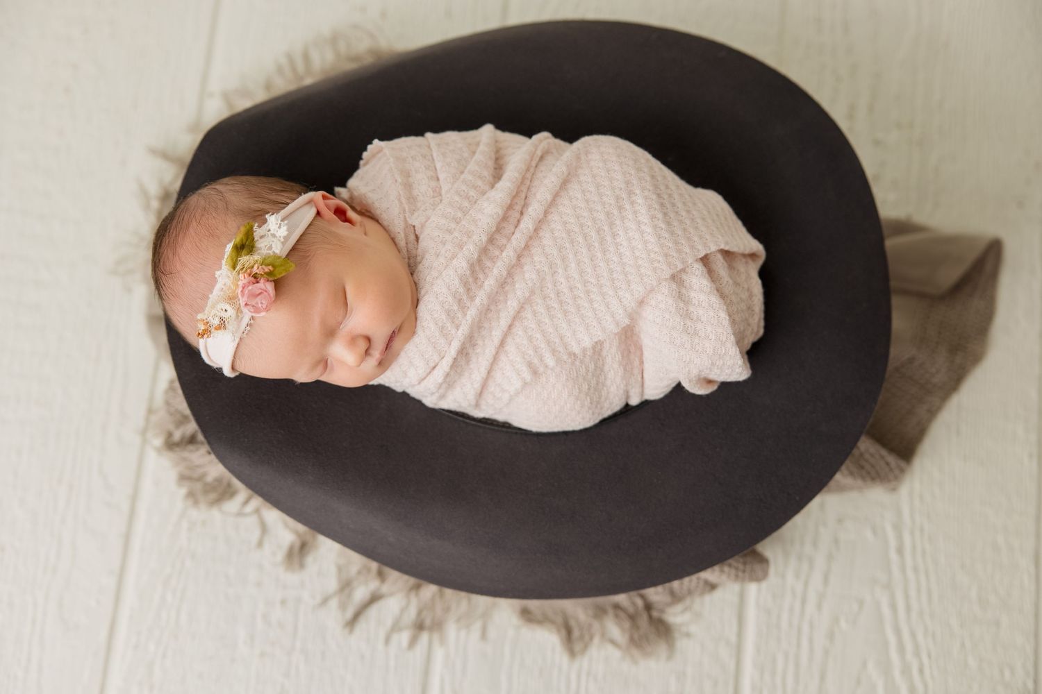 Sleeping newborn nestled in plush dark gray circular nursing pillow on white wooden floor.