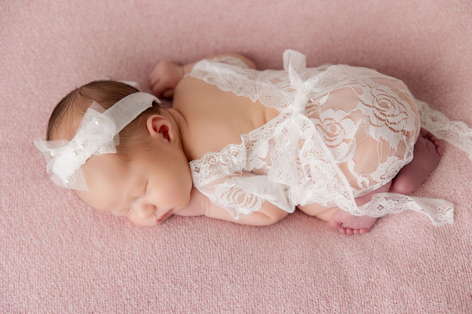 Peaceful sleeping newborn in white lace dress lying on pink textured backdrop.