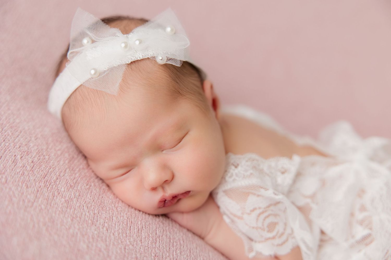 Close-up newborn photography showing baby in white lace on pink fabric backdrop.