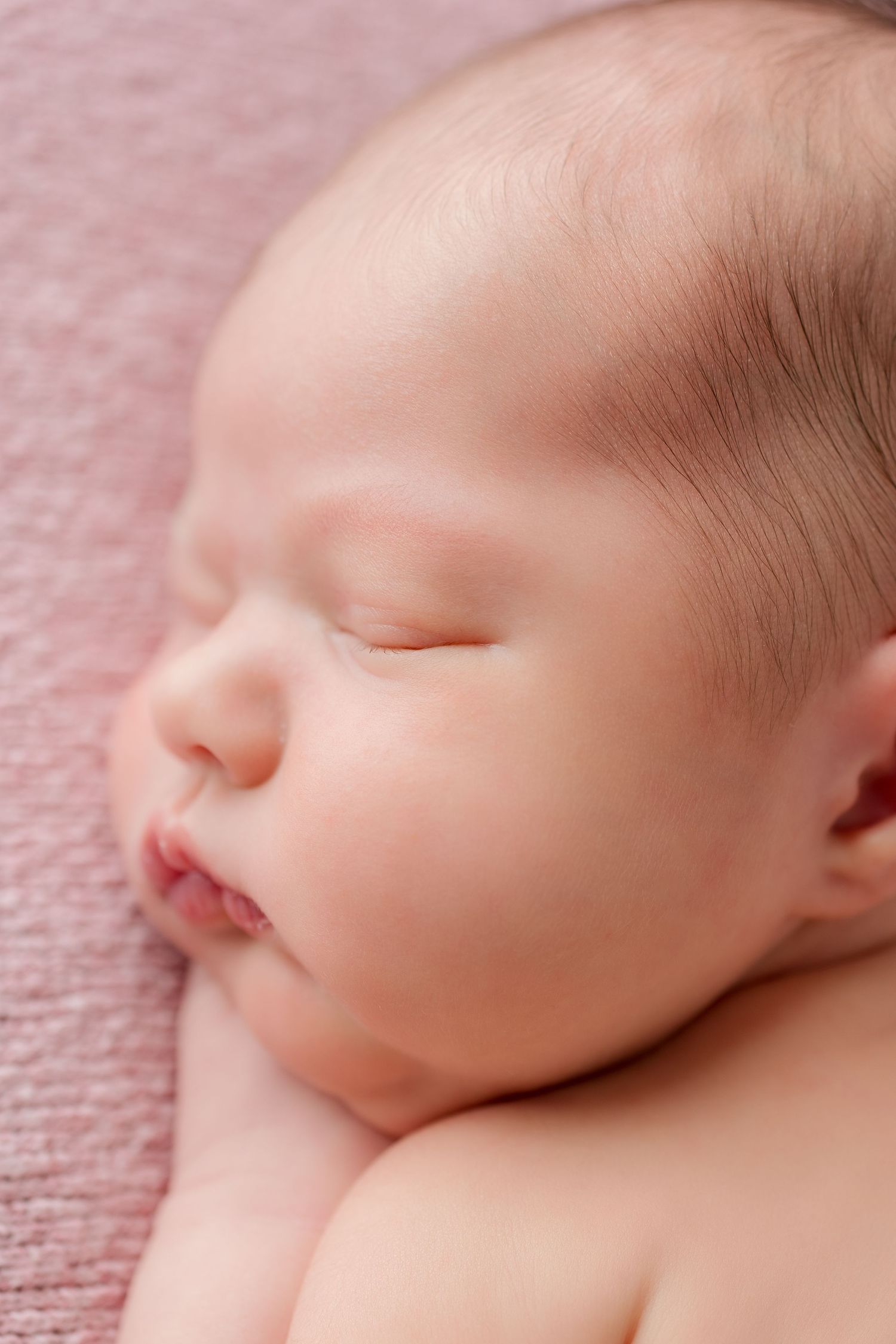 Gentle close-up macro shot of a sleeping newborn's face against a soft pink textured background.