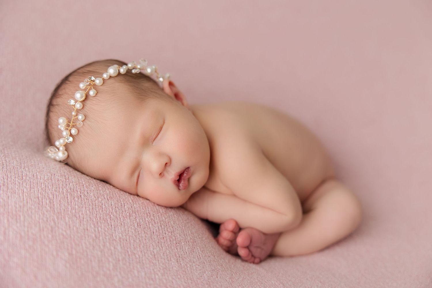 Artistic portrait of sleeping newborn baby posed on pink blanket with pearl headband accent.