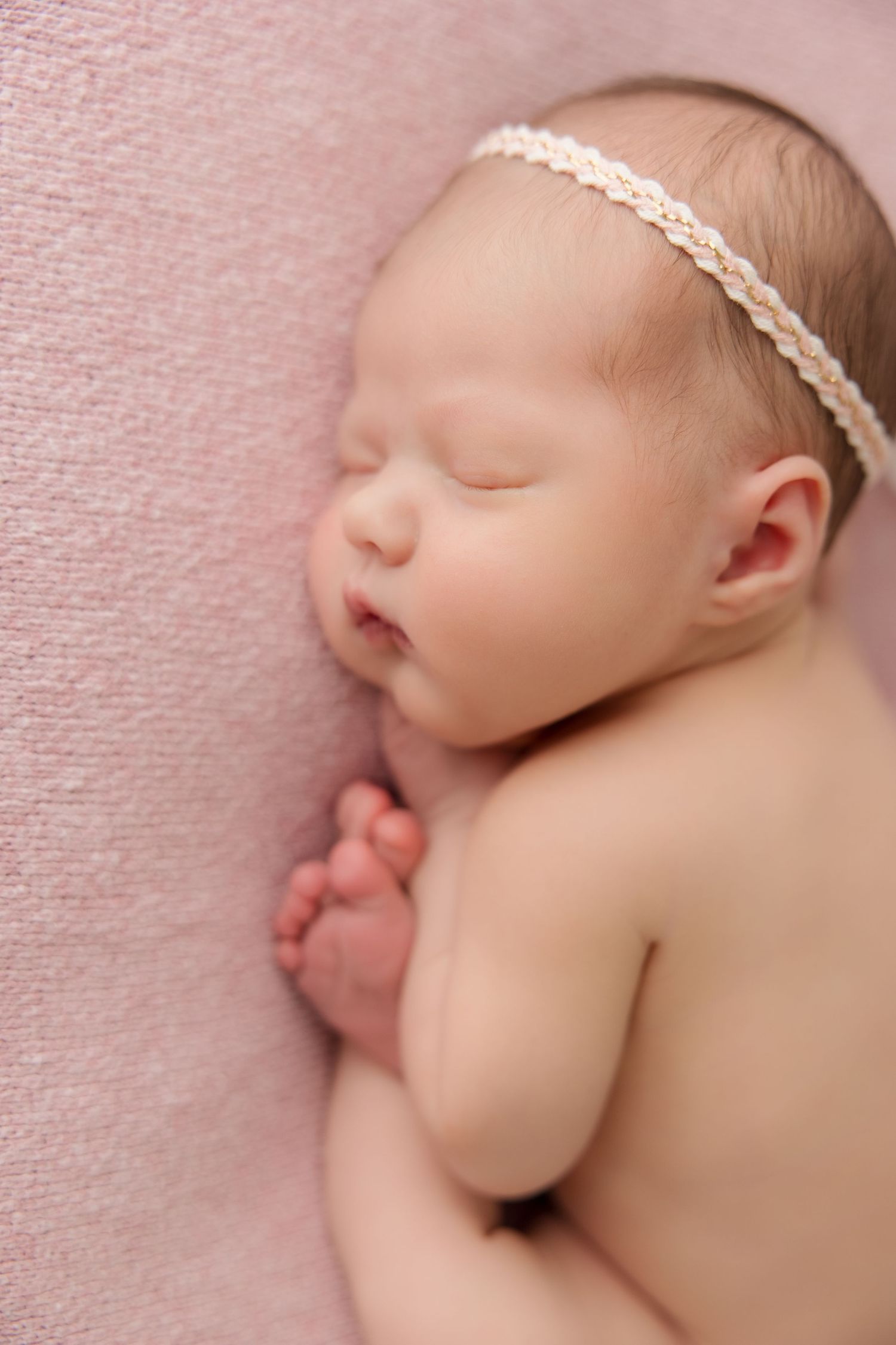 Newborn baby sleeping peacefully on pink blanket wearing delicate braided headband in artistic close-up portrait.