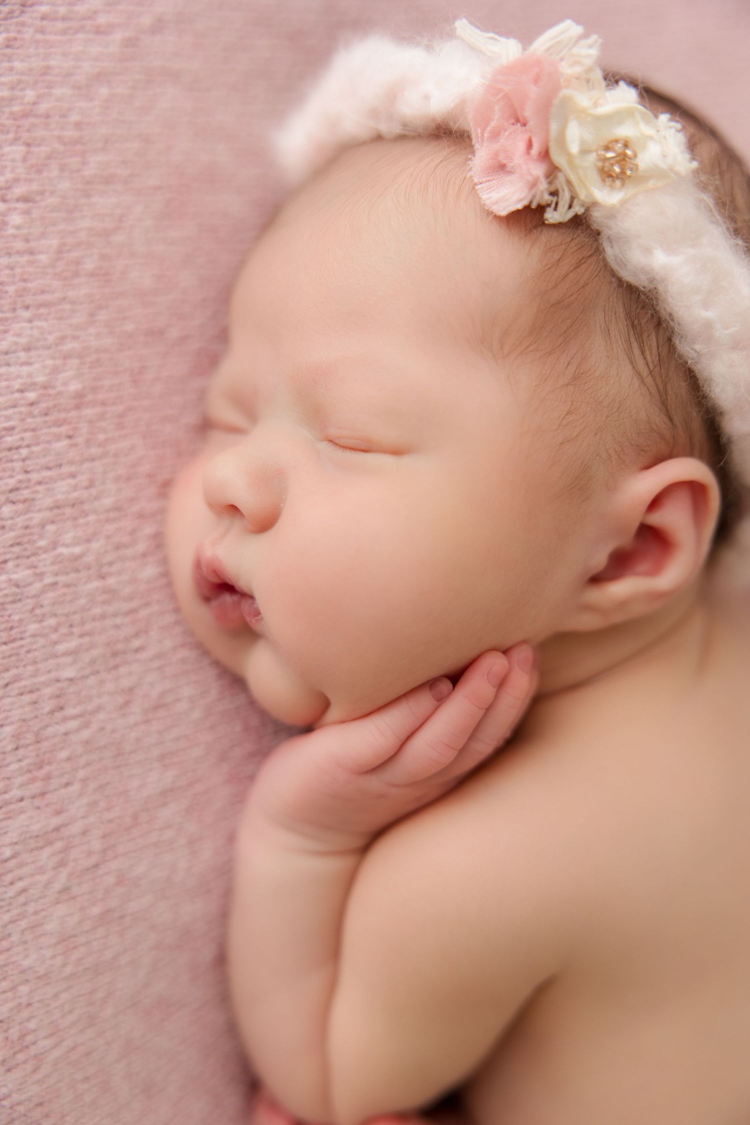 Close-up portrait of sleeping newborn baby with pink flower headband against soft pink textured background.