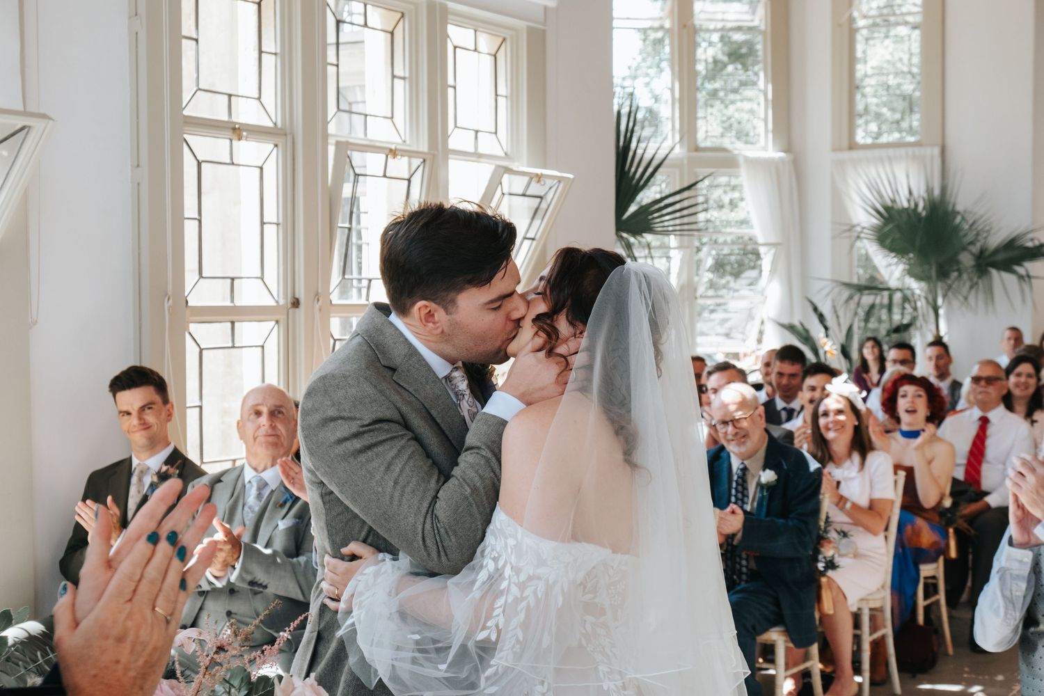 A bride and groom kiss at their wedding ceremony. Family and friends clap in the background as they kiss.