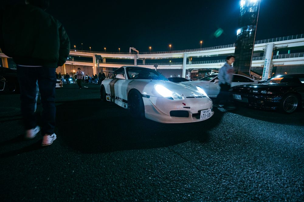 A nighttime scene showing several sports cars parked in a dark parking area with bright headlights illuminating the space.