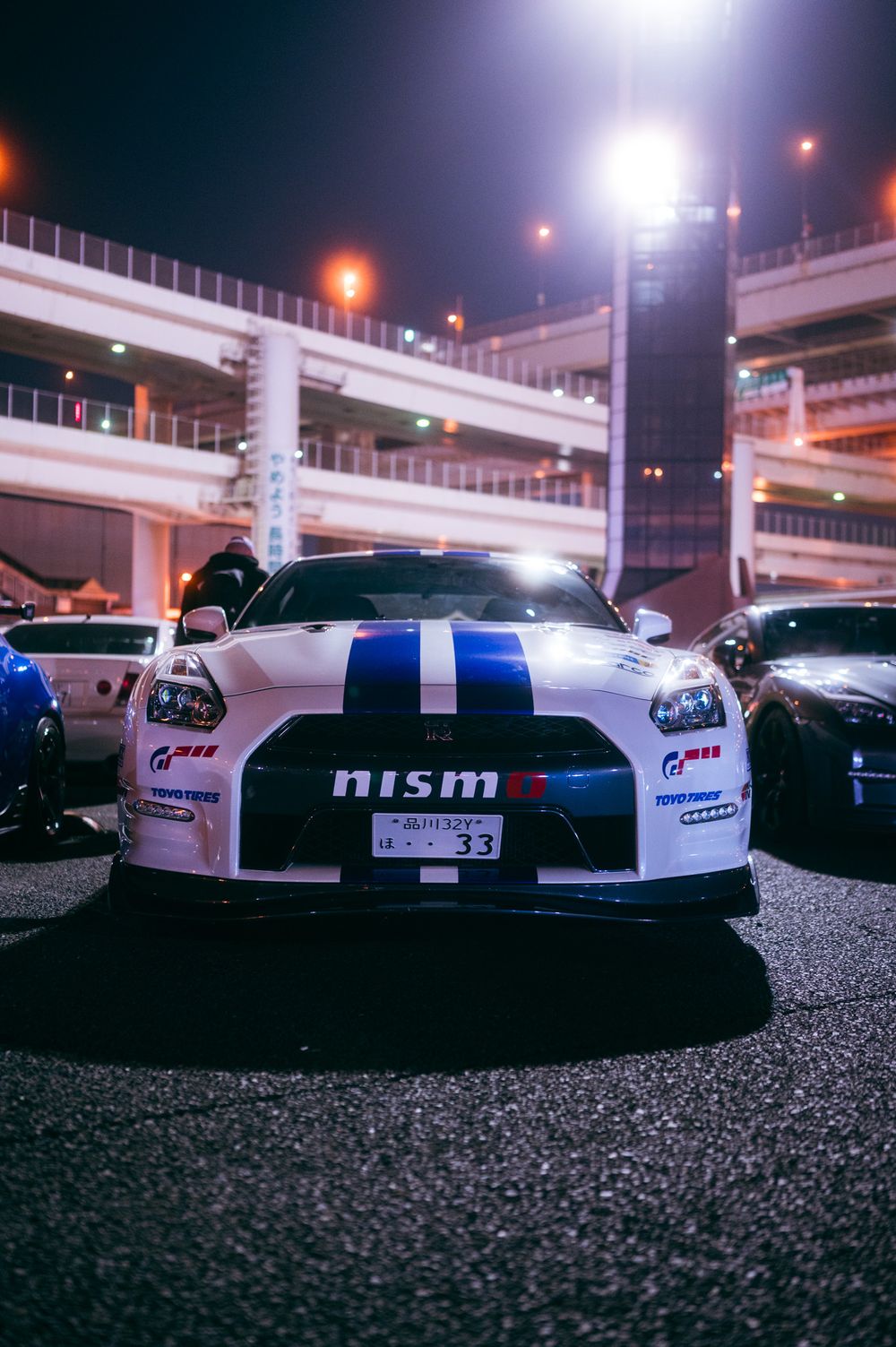 Low-angle view of white Nissan GT-R NISMO race cars with blue stripes under nighttime stadium lights.