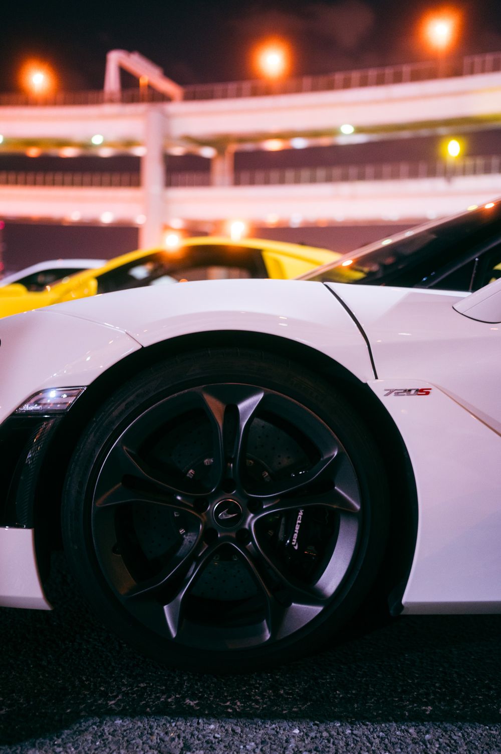 Close-up of a white sports car's black alloy wheel with orange city lights glowing in the background at night.