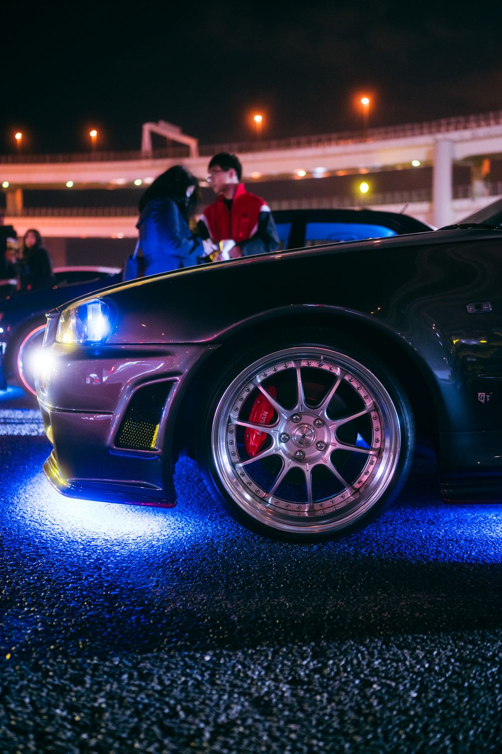 Low angle nighttime shots showing illuminated car wheels and undercarriage with blue lighting effects on dark pavement.