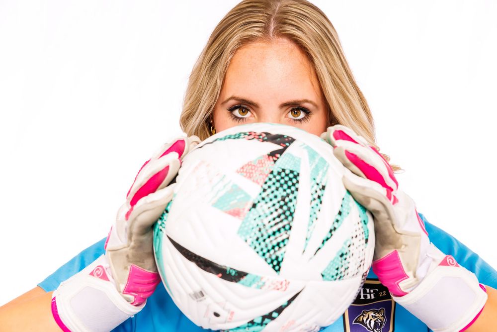 A soccer goalkeeper in blue jersey and pink gloves holds a white ball in front of their face.