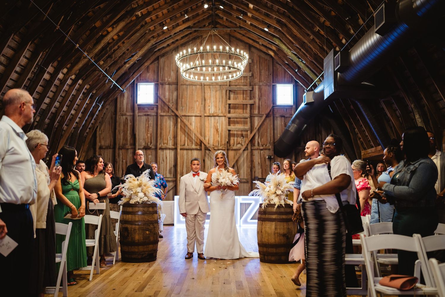 Wedding guests gather inside a rustic wooden barn with elegant chandeliers casting warm light over the celebration.