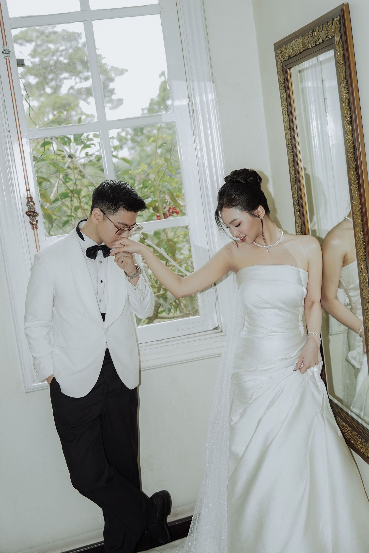 Groom in white tuxedo kisses brides hand as she stands beside a gilded mirror near a window.
