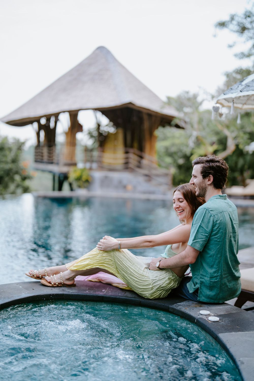 Couple relaxing by an infinity pool at a luxury tropical resort with thatched roof bungalows in the background.
