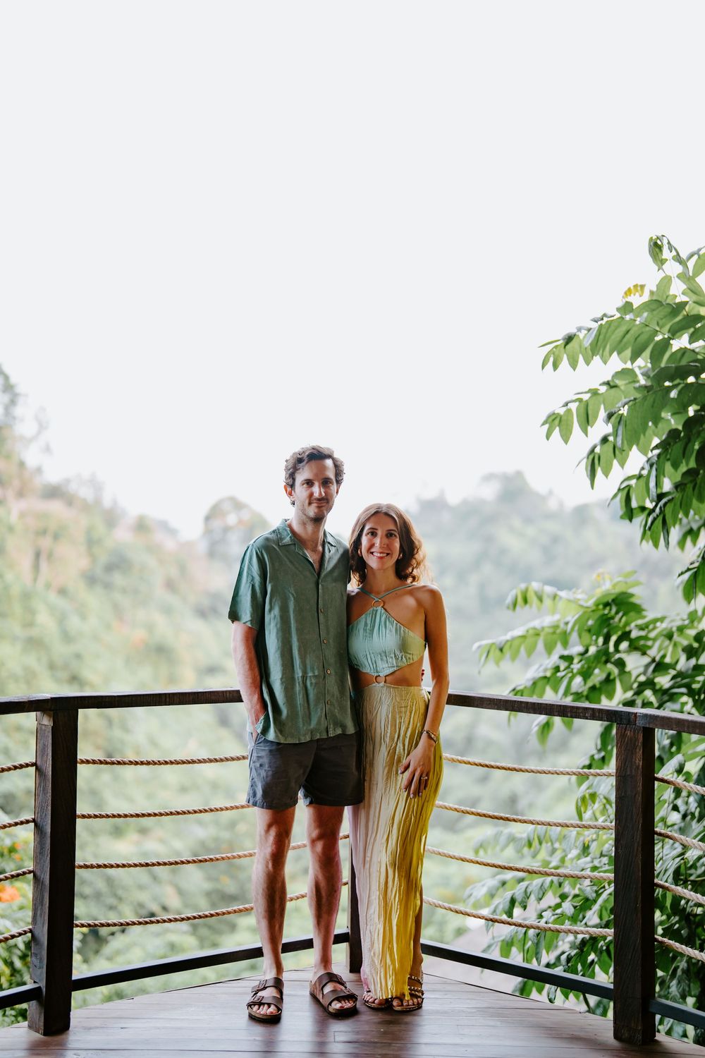 Couple standing on wooden deck with rope railing overlooking tropical mountain landscape.