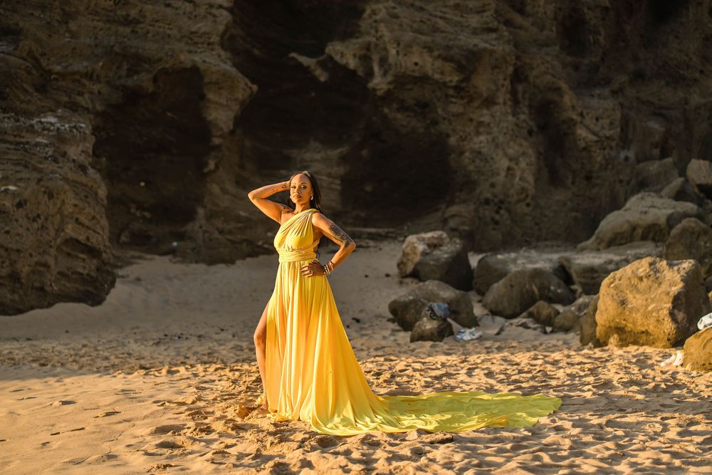 A woman poses in a sunny yellow gown on a sandy beach surrounded by dramatic rocky cliffs.