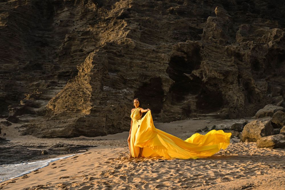Flowing yellow dress billows dramatically against rocky coastal cliffs during golden hour photography session on beach.